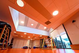 Salle motricité orange, structures bois, luminaires circulaires encastrés, plafond géométrique et éclairage chaud.