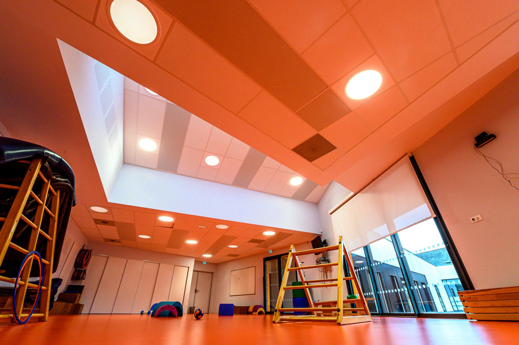 Salle motricité orange, structures bois, luminaires circulaires encastrés, plafond géométrique et éclairage chaud.