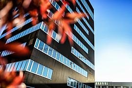 Façade sombre du bâtiment Condate à Rennes avec larges fenêtres horizontales sous un ciel bleu et feuillage rouge flou