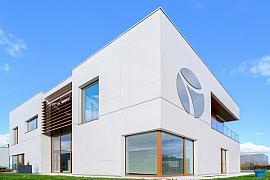 Façade béton blanc bâtiment moderne, menuiseries bois, logo circulaire alu sous un ciel bleu dégagé 