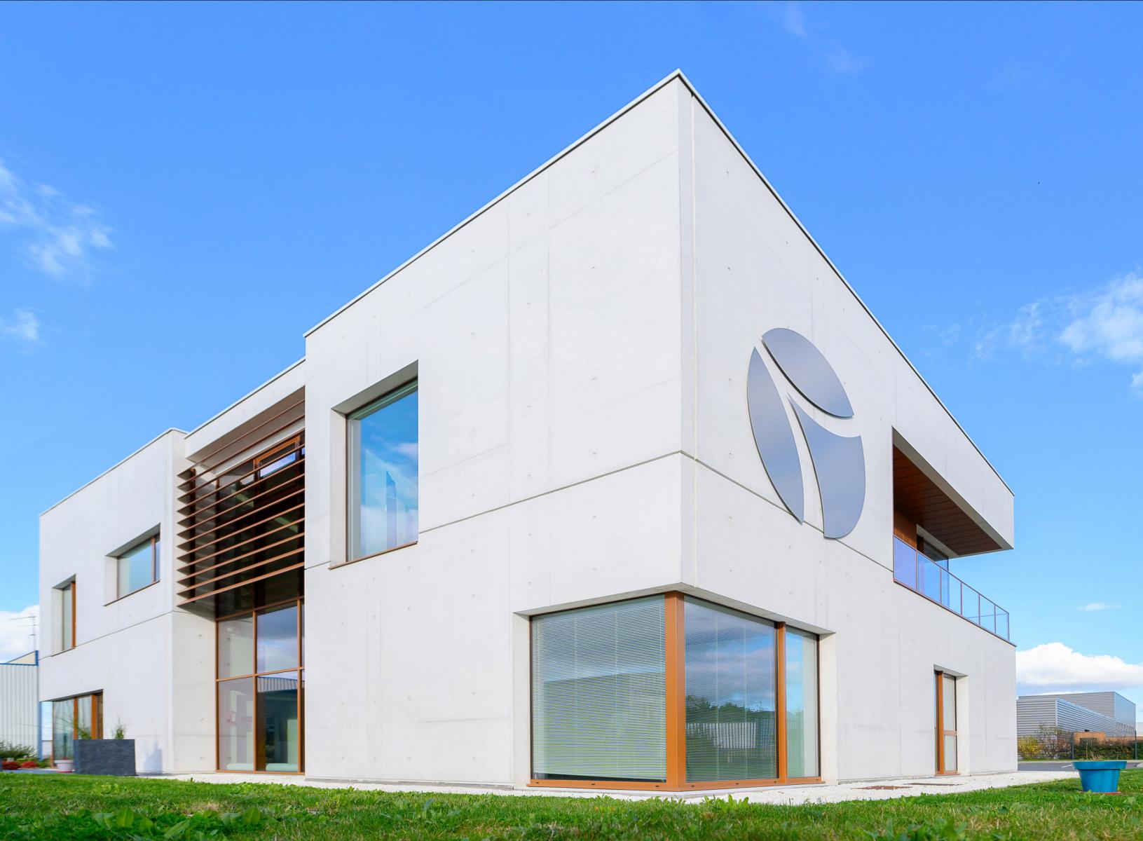 Façade béton blanc bâtiment moderne, menuiseries bois, logo circulaire alu sous un ciel bleu dégagé 