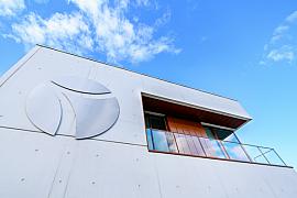 Contre-plongée d’une façade moderne en béton blanc avec balcon en bois et logo minimaliste sous ciel bleu.