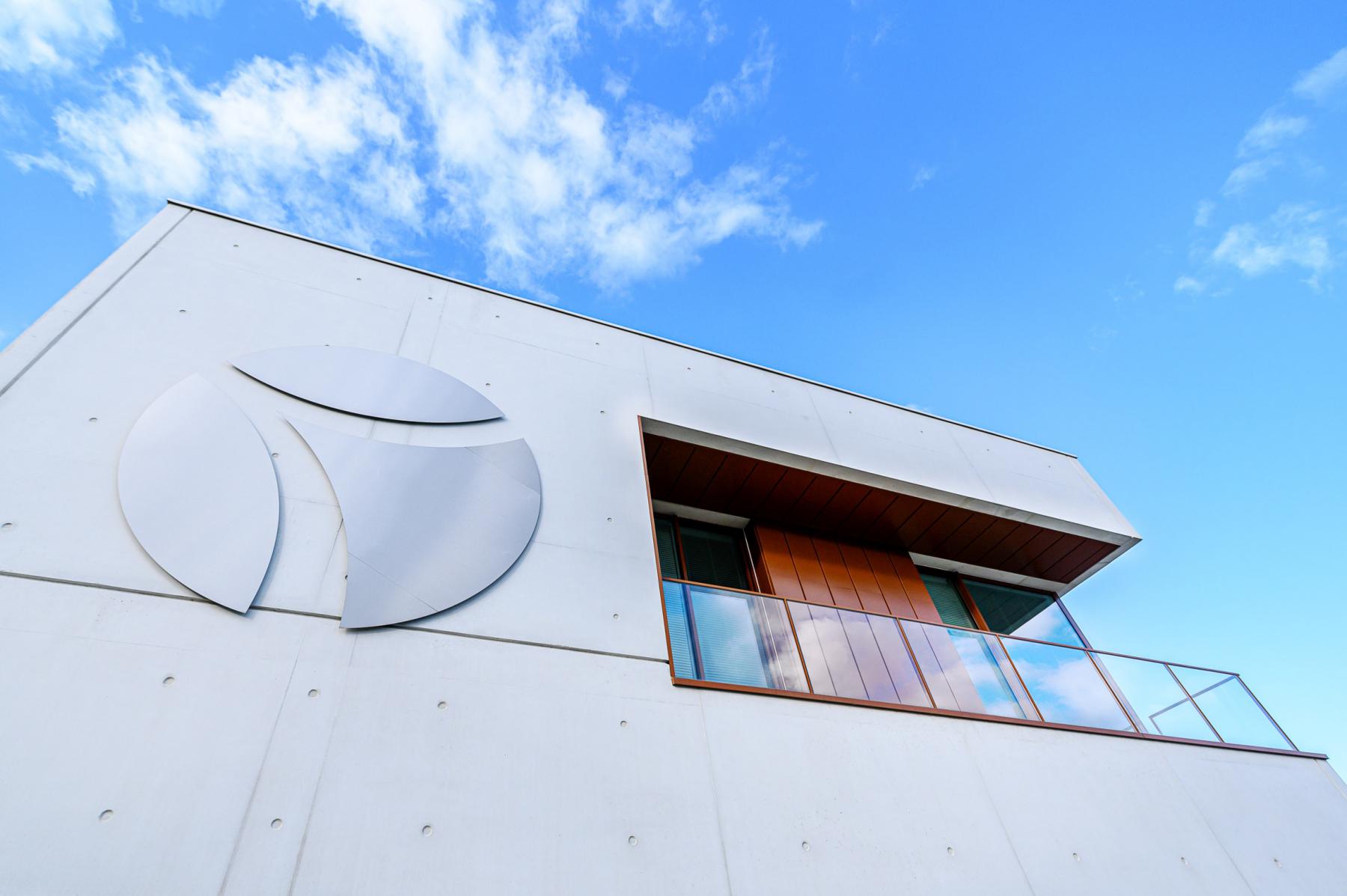 Contre-plongée d’une façade moderne en béton blanc avec balcon en bois et logo minimaliste sous ciel bleu.