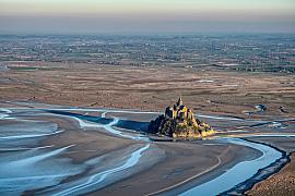 Abbaye du Mont Saint-Michel isolée sur le sable à marée basse avec des chenaux d’eau sinueux sous un ciel clair.
