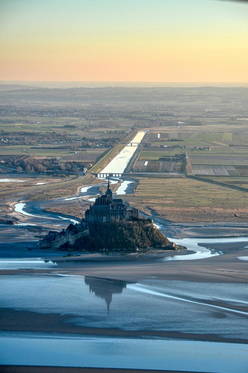 Vue aérienne du Mont Saint-Michel et du Couesnon serpentant entre les bancs de sable sous une lumière de fin de journée.