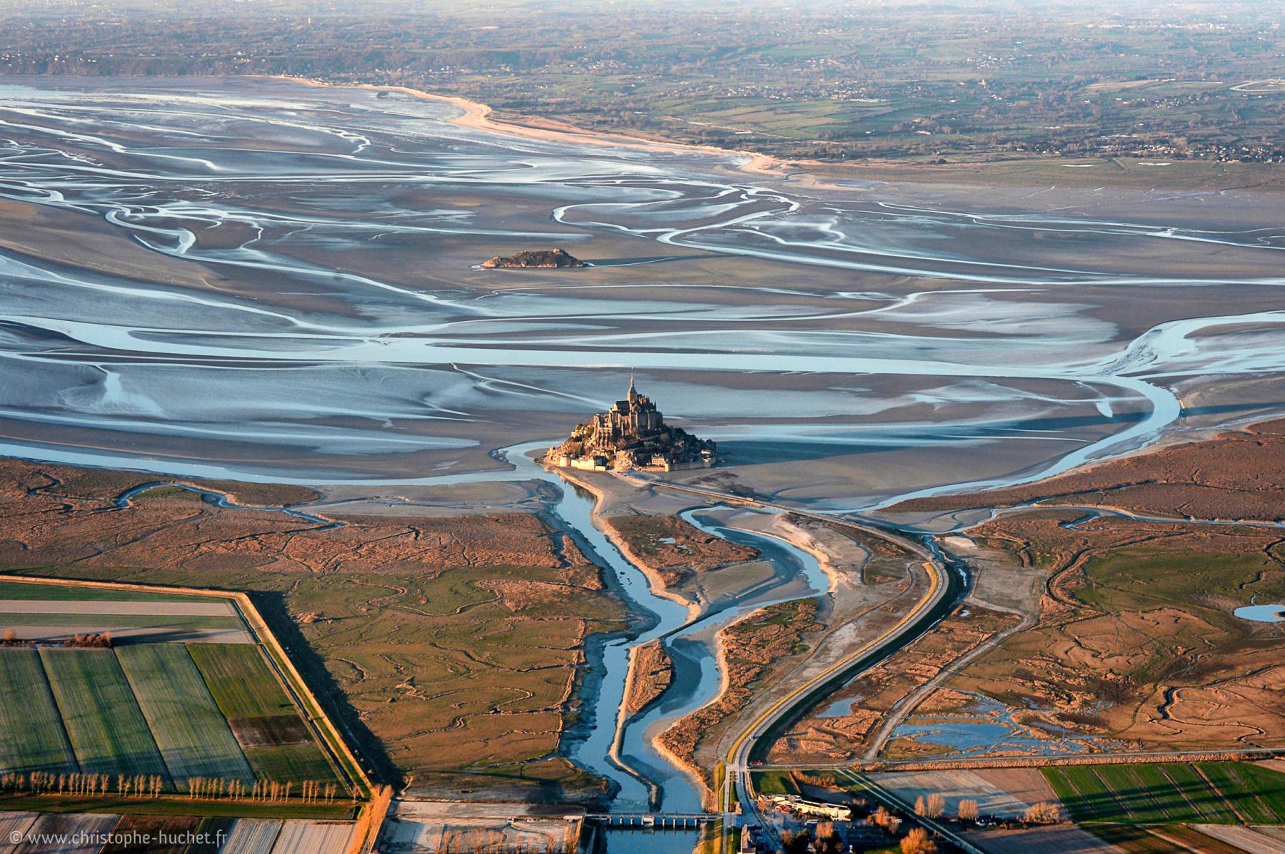 Vue aérienne du Mont Saint-Michel entouré de chenaux d’eau à marée basse et de polders verdoyants en avant-plan.