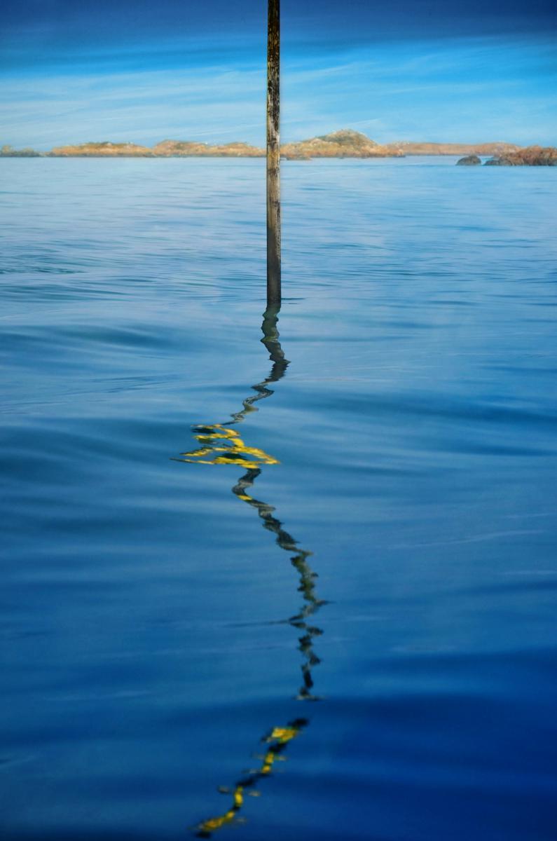 Balise maritime verticale et son reflet jaune sur une mer bleue calme face aux îles de l’archipel de Chausey.
