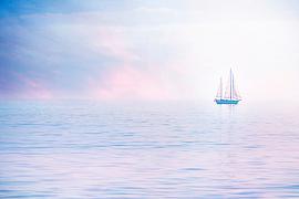 Voilier à deux mâts naviguant sur une mer calme enveloppée d’une brume aux teintes pastel roses et bleues à Chausey.