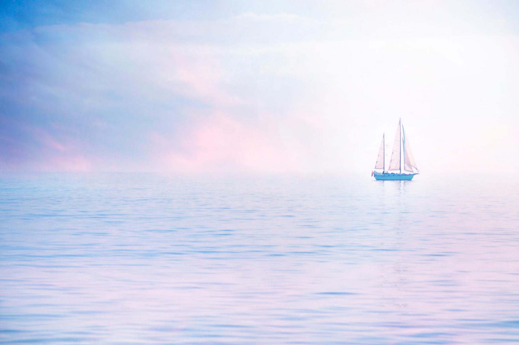 Voilier à deux mâts naviguant sur une mer calme enveloppée d’une brume aux teintes pastel roses et bleues à Chausey.