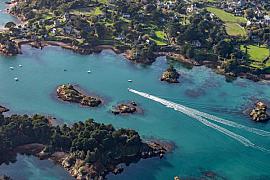 Vue du ciel des eaux turquoise de Bréhat avec le sillage blanc d’un bateau naviguant entre les îlots rocheux boisés.