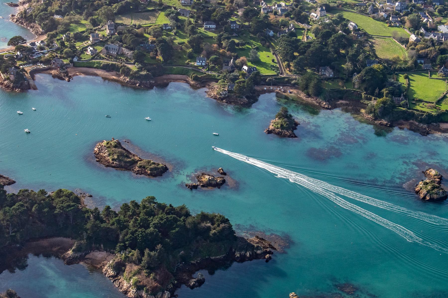 Vue du ciel des eaux turquoise de Bréhat avec le sillage blanc d’un bateau naviguant entre les îlots rocheux boisés.