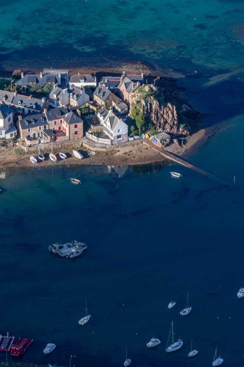 Maisons de pierre au bord de l’eau turquoise avec des bateaux au mouillage dans le port breton de Loguivy sous une lumière zénithale.