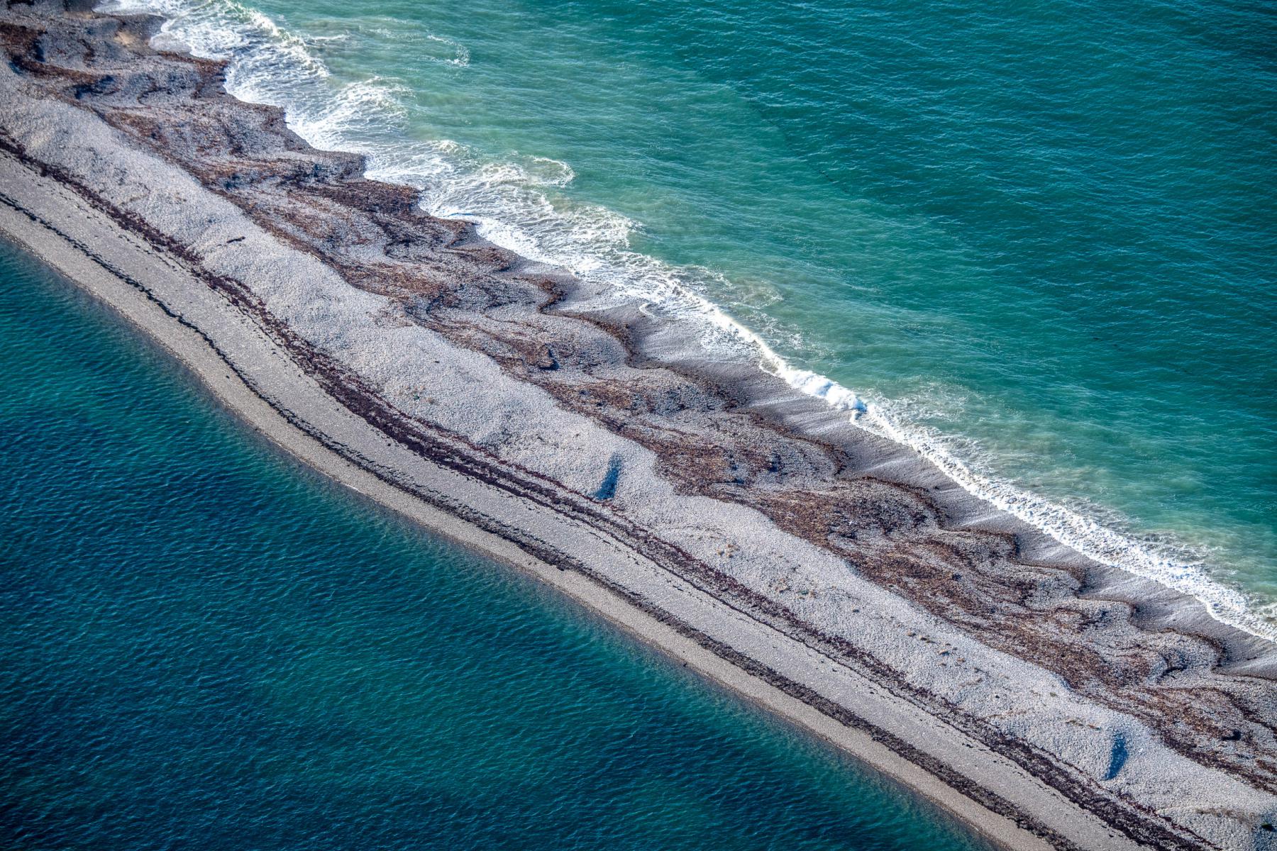 Vue aérienne d’une flèche de galets gris bordée par une mer turquoise et de fines vagues blanches à Pleubian en Bretagne.