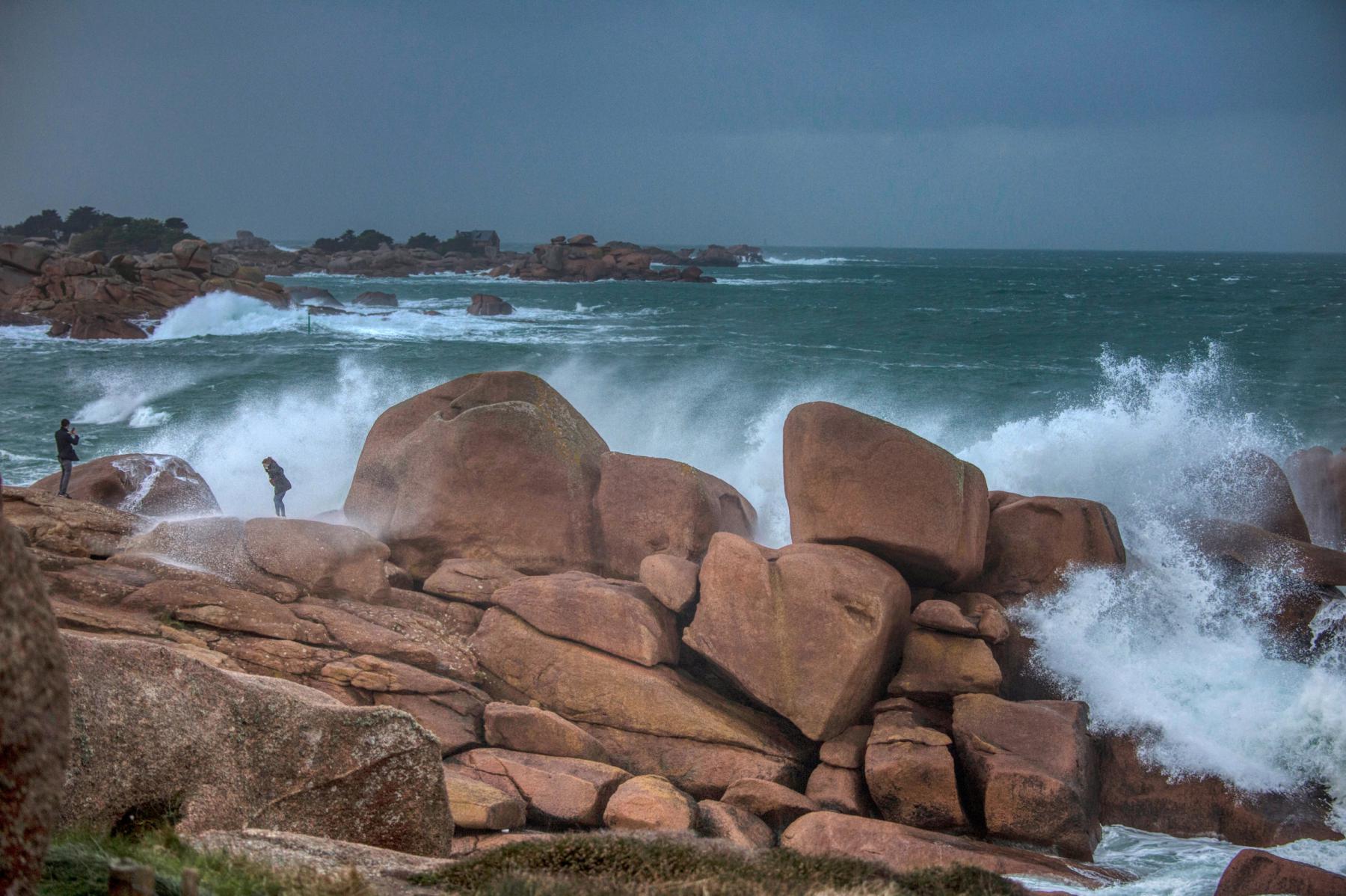 Vagues puissantes s’écrasant sur les rochers de Ploumanac’h sous un ciel sombre de tempête avec silhouettes de promeneurs.
