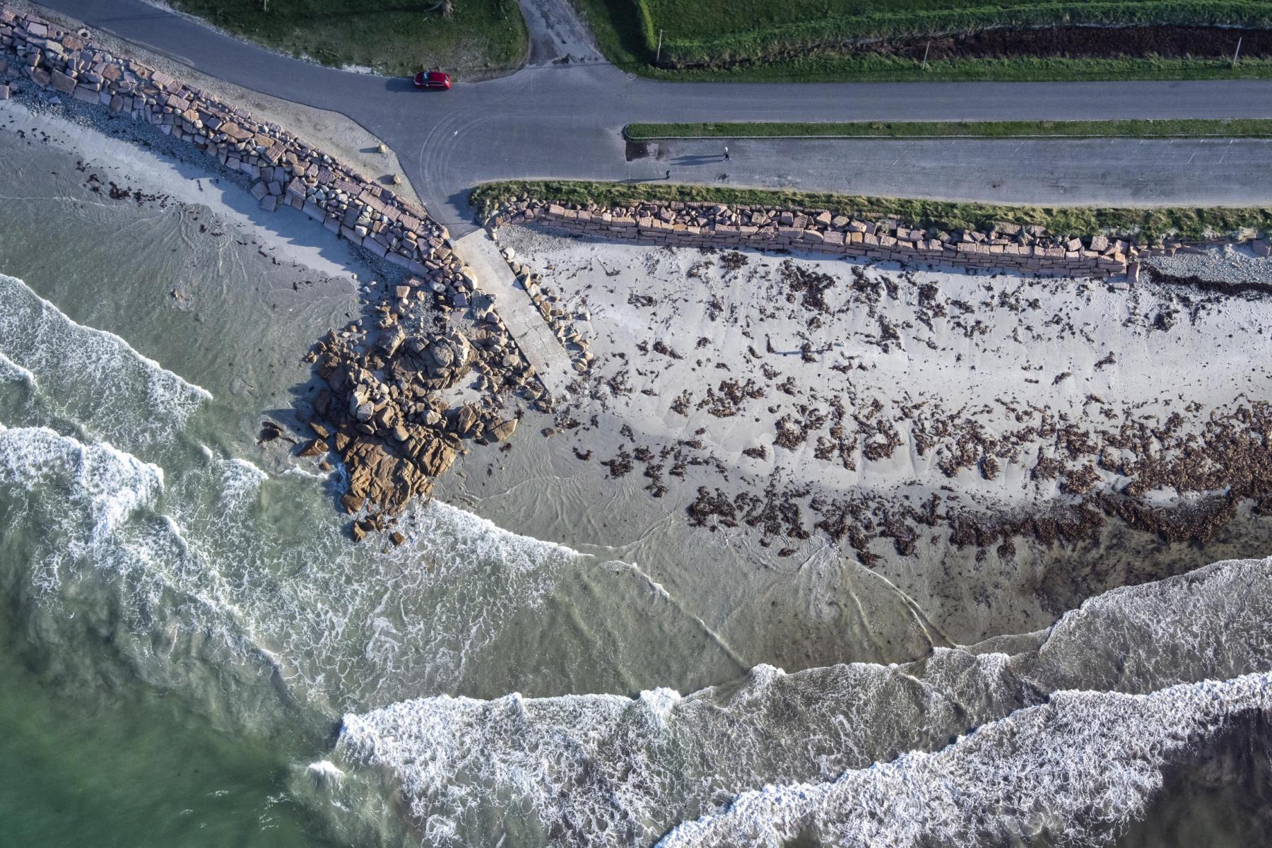 Vue aérienne verticale d’une plage de sable blanc avec vagues écumantes, rochers et route littorale à Trévou-Tréguignec.