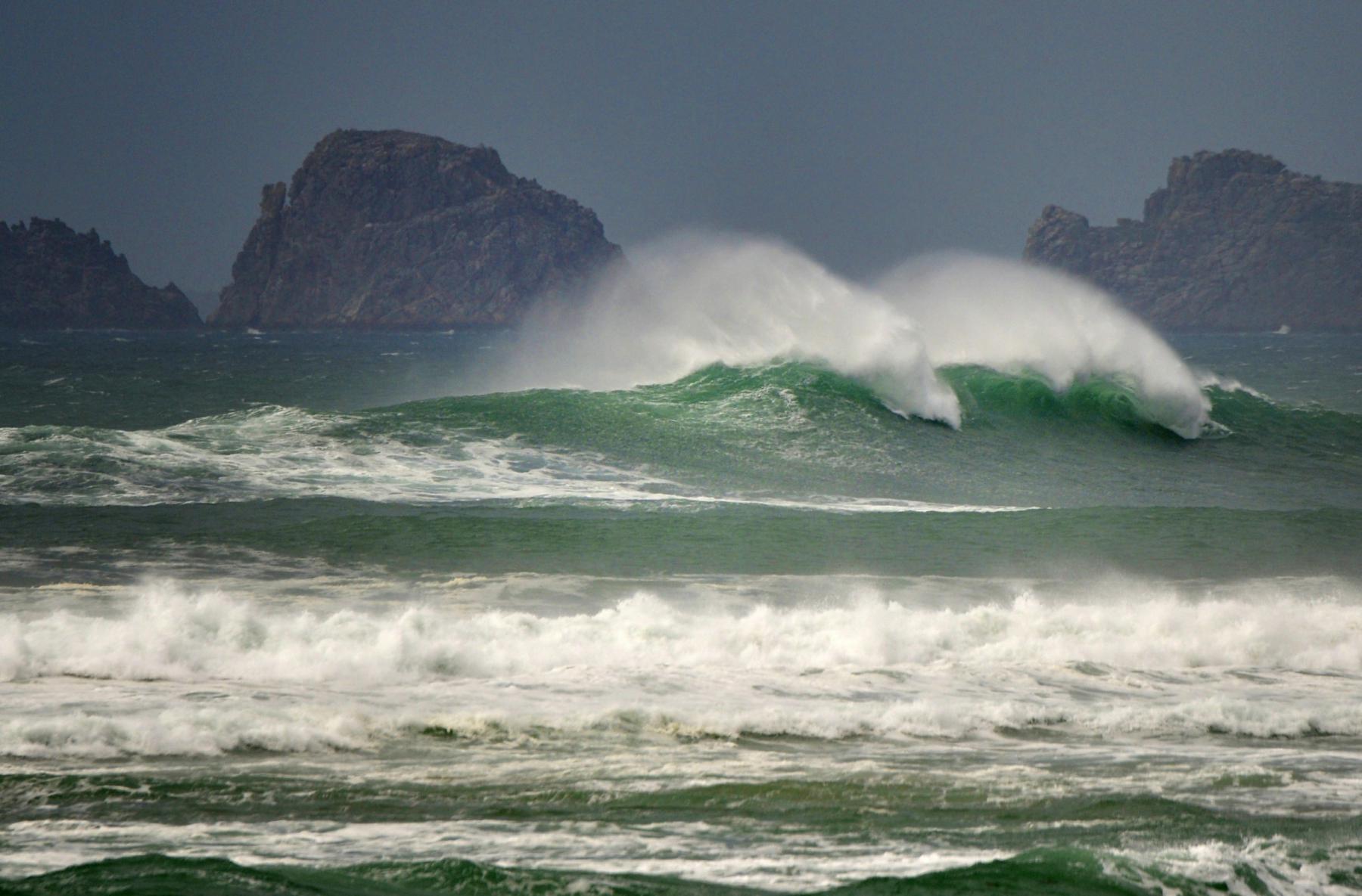 Écume blanche et vagues vert émeraude déferlant devant des falaises rocheuses sombres sous un ciel de tempête en Bretagne.