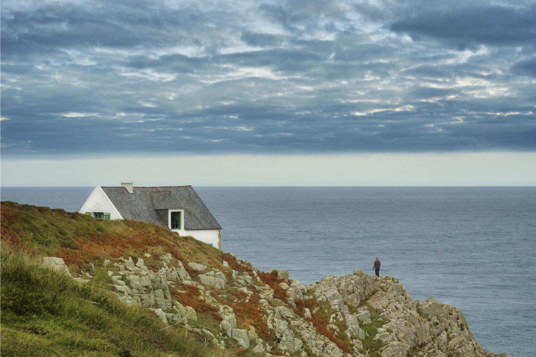 Petite maison blanche isolée sur une falaise rocheuse à la pointe de Bac’haol avec uen silhouette posée sur les rochers sous un ciel gris et glaz