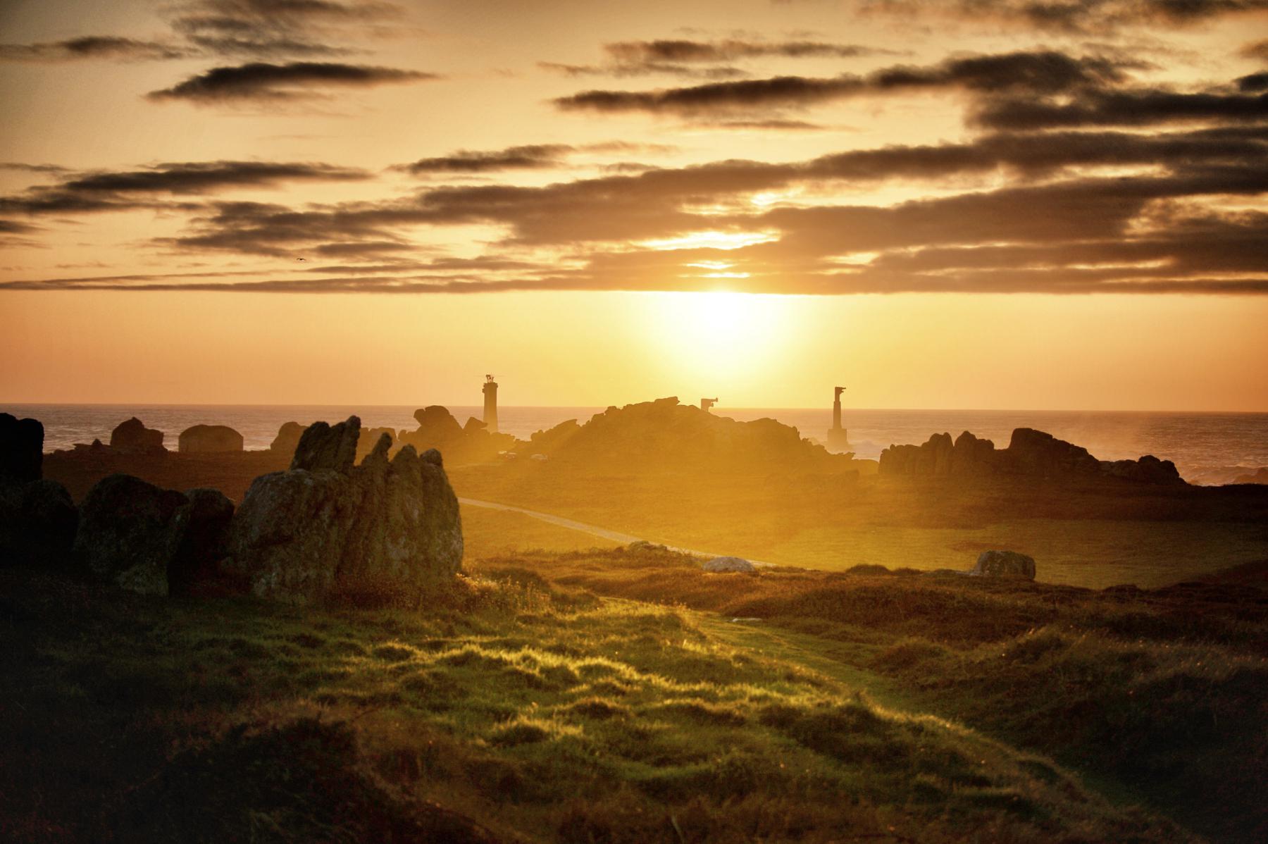 Soleil couchant doré illuminant les rochers de la pointe de Pern à Ouessant, silhouette de phares à l’horizon sous un ciel nuageux.