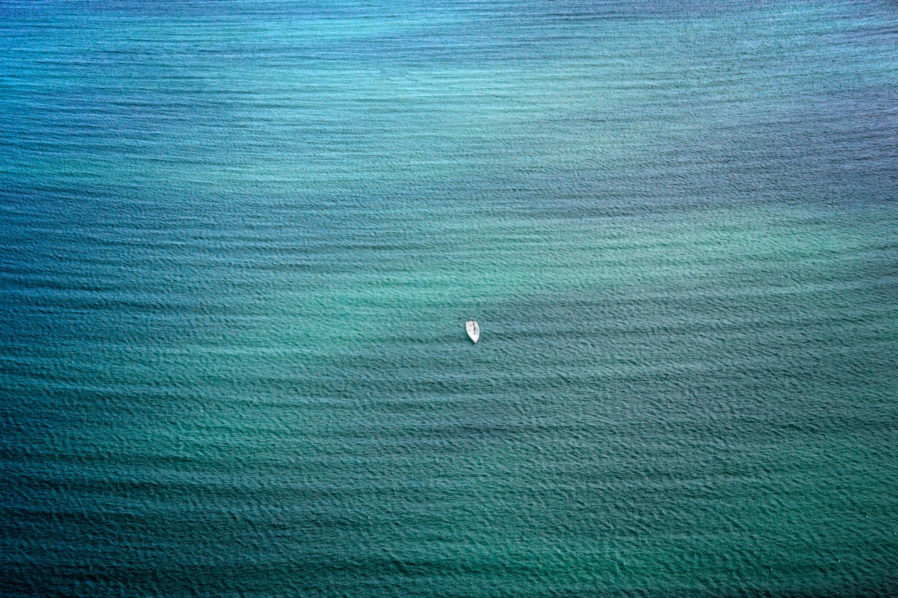 Vue de dessus d’un petit bateau blanc isolé sur une mer aux dégradés de bleu turquoise et de vert émeraude en Bretagne.