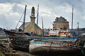 Épaves de bateaux en bois rouillés au premier plan devant la chapelle de Rocamadour et la tour Vauban sous un ciel nuageux.