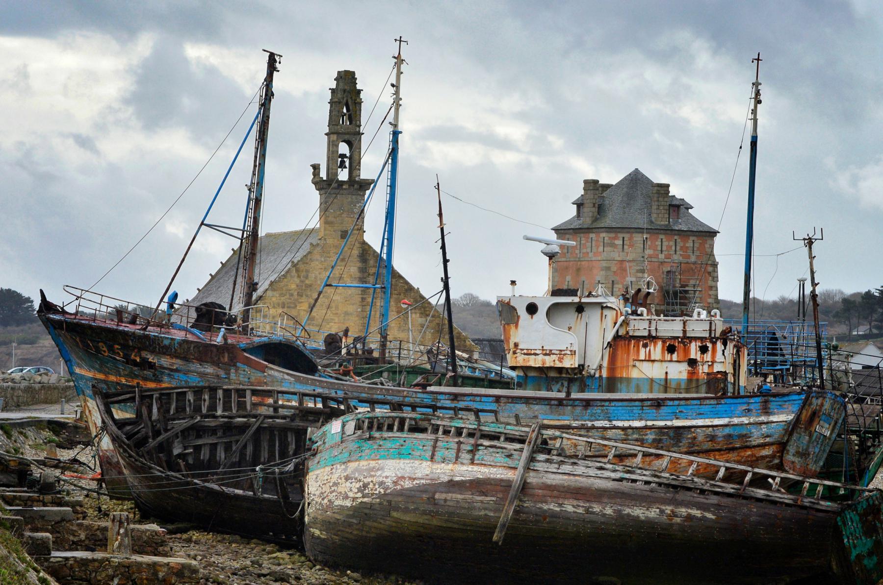 Épaves de bateaux en bois rouillés au premier plan devant la chapelle de Rocamadour et la tour Vauban sous un ciel nuageux.