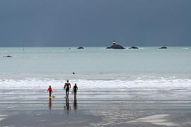 Trois surfeurs marchant sur le sable humide vers les vagues sous un ciel de tempête à Saint-Lunaire en Bretagne.