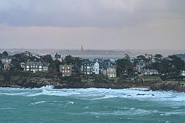 Vagues d’écume blanche se brisant contre les rochers sous des villas bretonnes et un ciel de tempête gris.