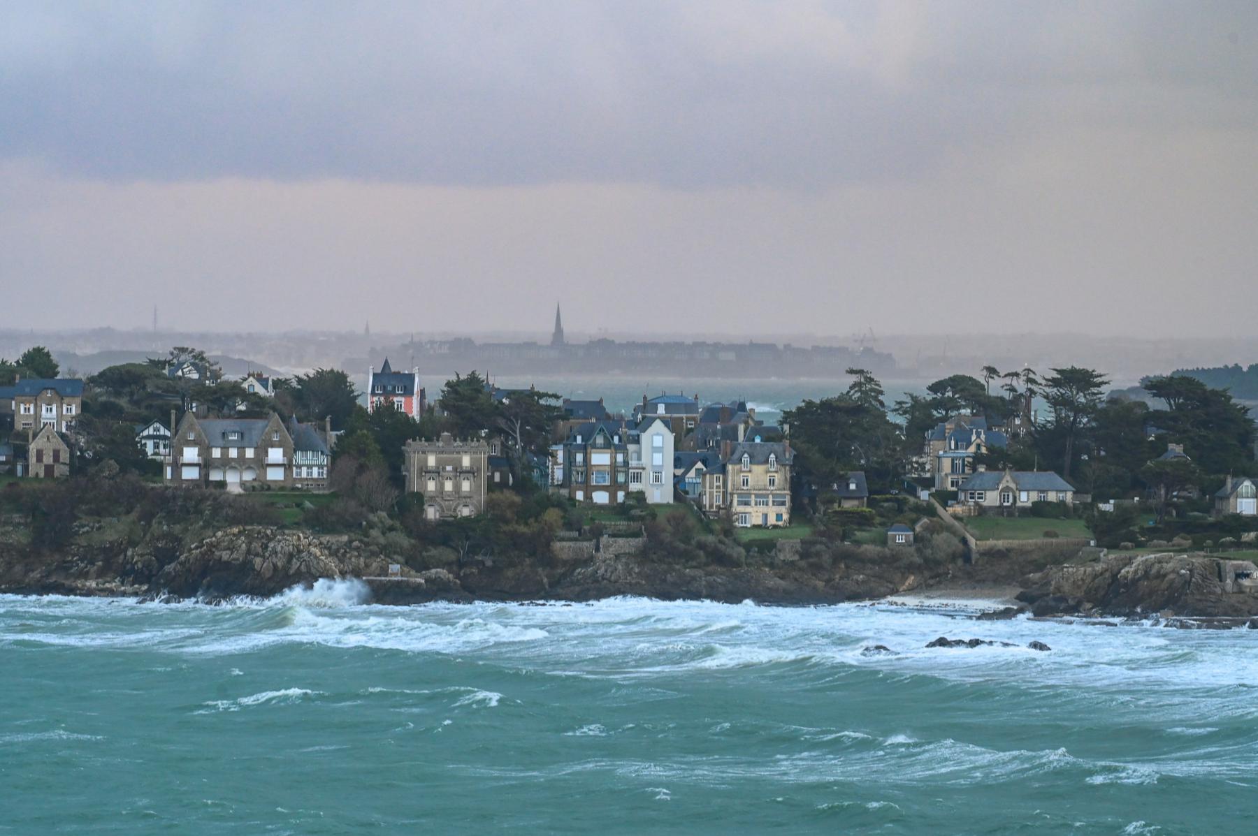 Vagues d’écume blanche se brisant contre les rochers sous des villas bretonnes et un ciel de tempête gris.