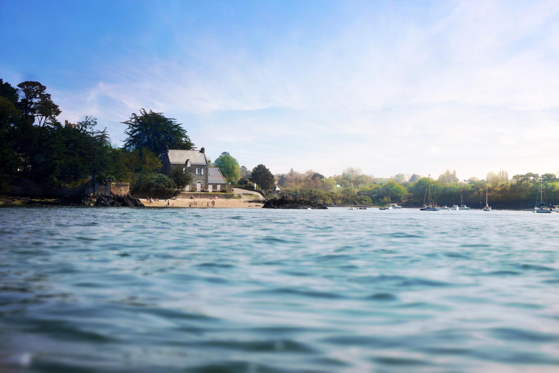 Maison en pierre sur les rives de la Rance à Saint-Malo, eau calme au premier plan et ciel azur.