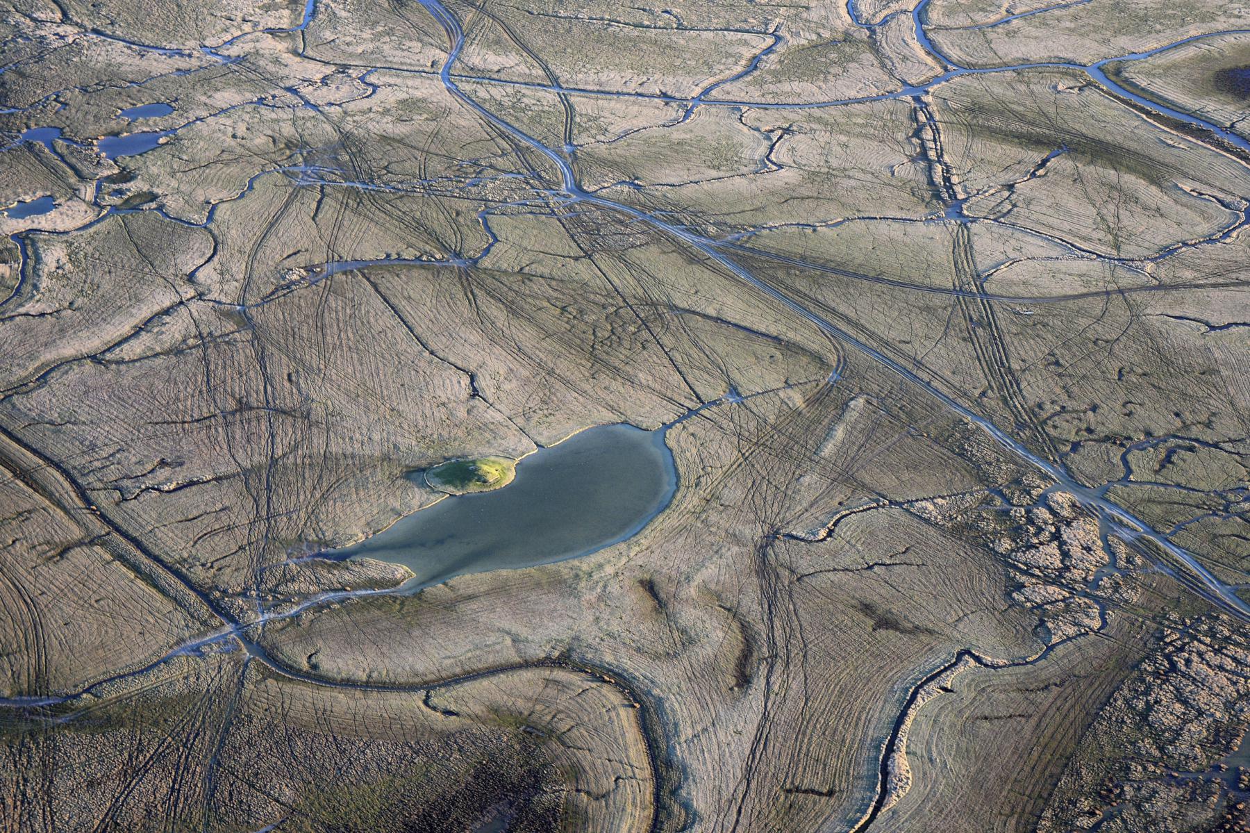 Vue aérienne de polders avec une mare de gabion entourée de chenaux d’eau et de traces dans la vase grise et beige.
