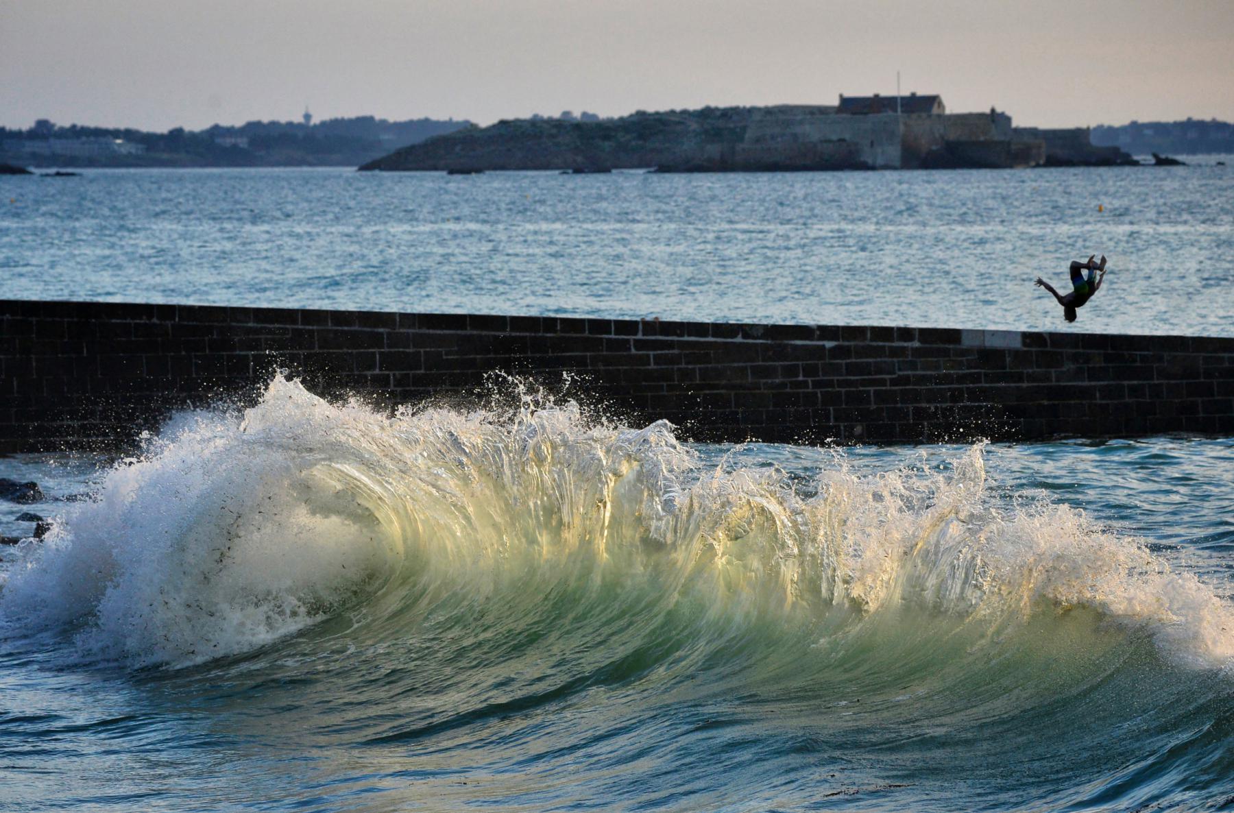 Écume d’une vague rétro-éclairée devant la digue de Saint-Malo avec la silhouette d’un homme faisant un salto arrière.