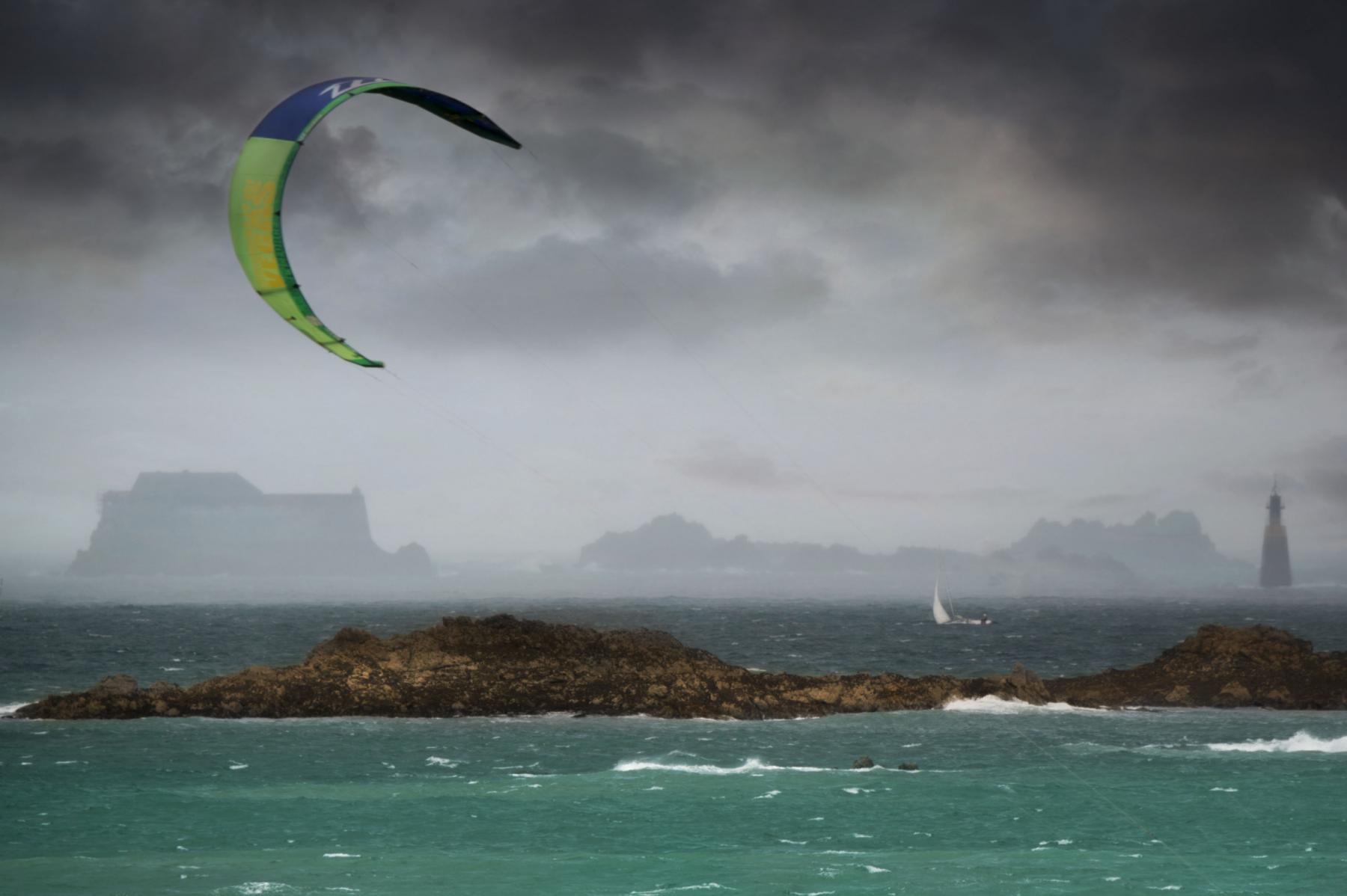 Voile de kitesurf verte dans un ciel orageux gris au-dessus d’une mer agitée avec le fort de la Conchée au loin