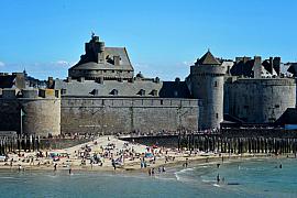 Les remparts du château de Saint-Malo surplombent une plage de sable bondée de monde sous un ciel bleu d’été.