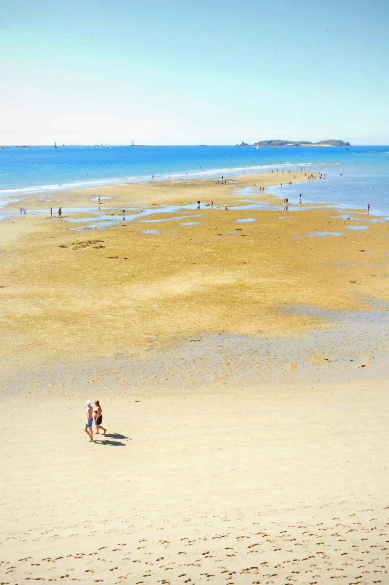 Promeneurs à marée basse, sur une vaste étendue de sable, mer turquoise et ciel bleu limpide à Saint-Malo.