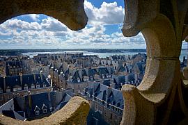 Toits d’ardoise de Saint-Malo vus à travers une ouverture en pierre sculptée de la cathédrale sous un ciel nuageux.