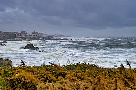 Vagues écumeuses se fracassant contre les rochers du Croisic sous un ciel gris de tempête avec végétation au premier plan.