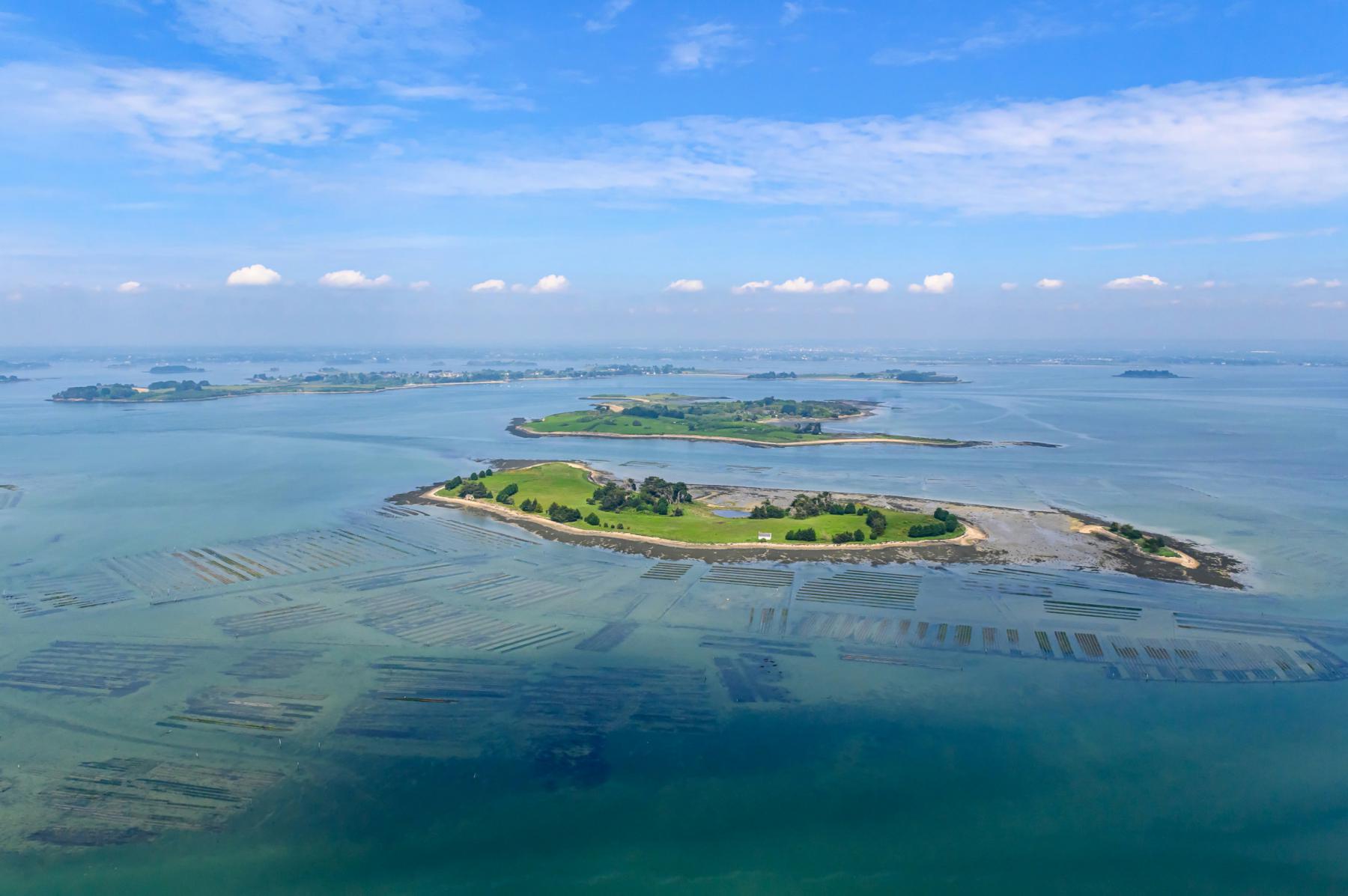 Vue du ciel des îles Ilur et d’Arz entourées d’eaux turquoise révélant des parcs à huîtres sous un ciel bleu d’été.