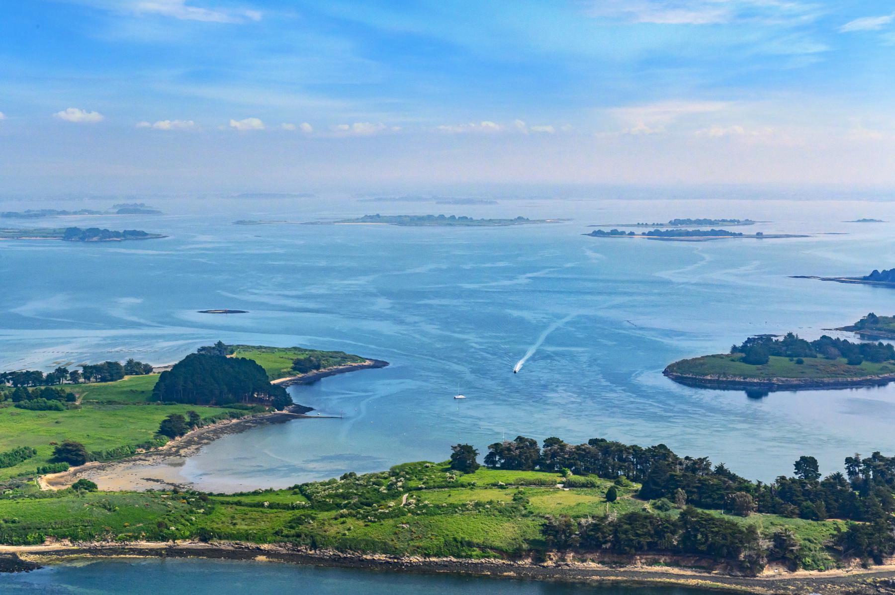Vue en plongée d’îlots verdoyants entourés d’une eau turquoise avec des sillages de bateaux sous un ciel clair.