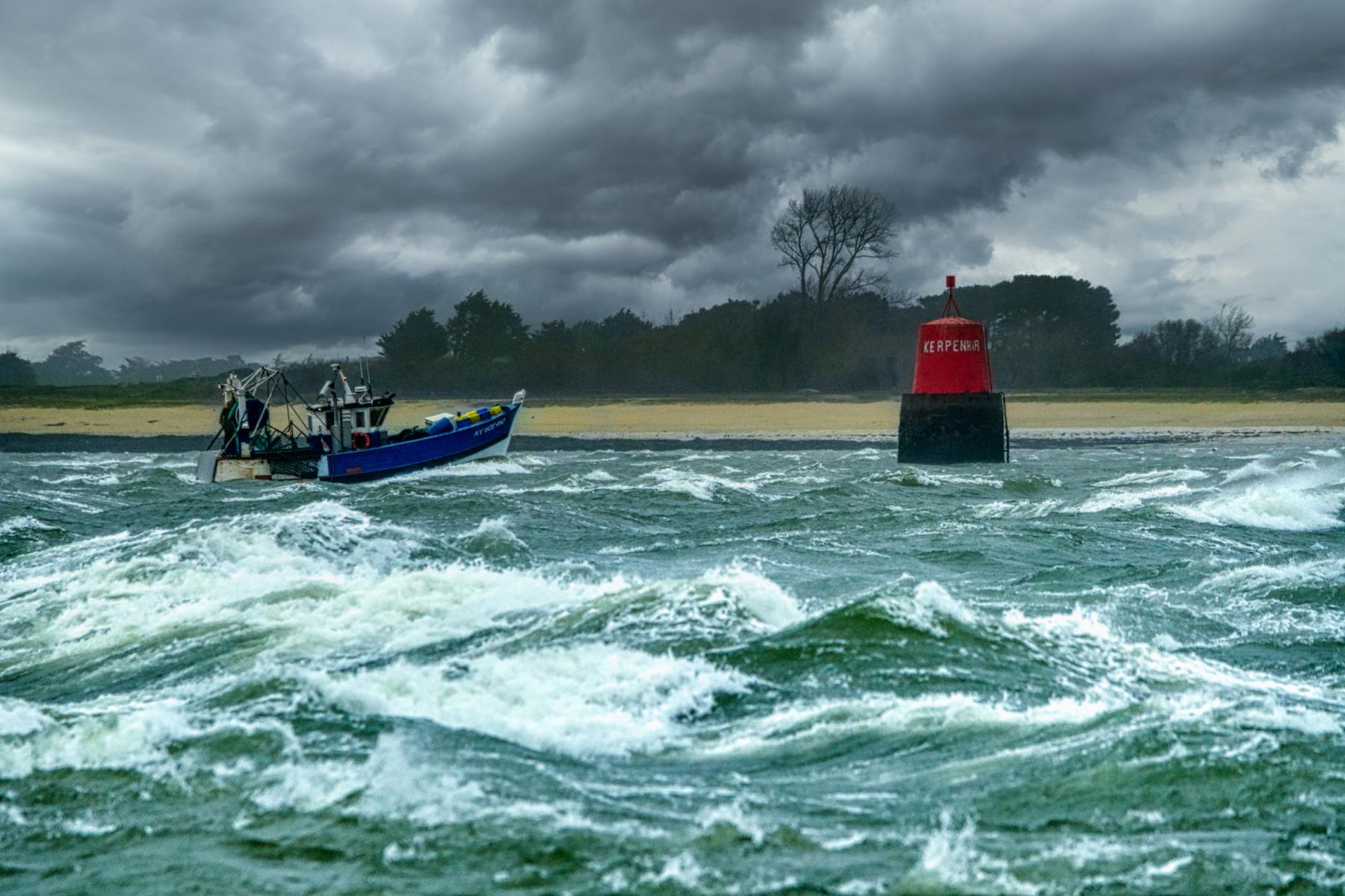 Bateau de pêche bleu luttant contre des vagues écumantes devant la balise rouge de Kerpenhir sous un ciel d’orage gris.