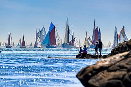 Voiliers traditionnels aux voiles colorées naviguent sur une mer bleue face à des spectateurs assis sur des rochers.