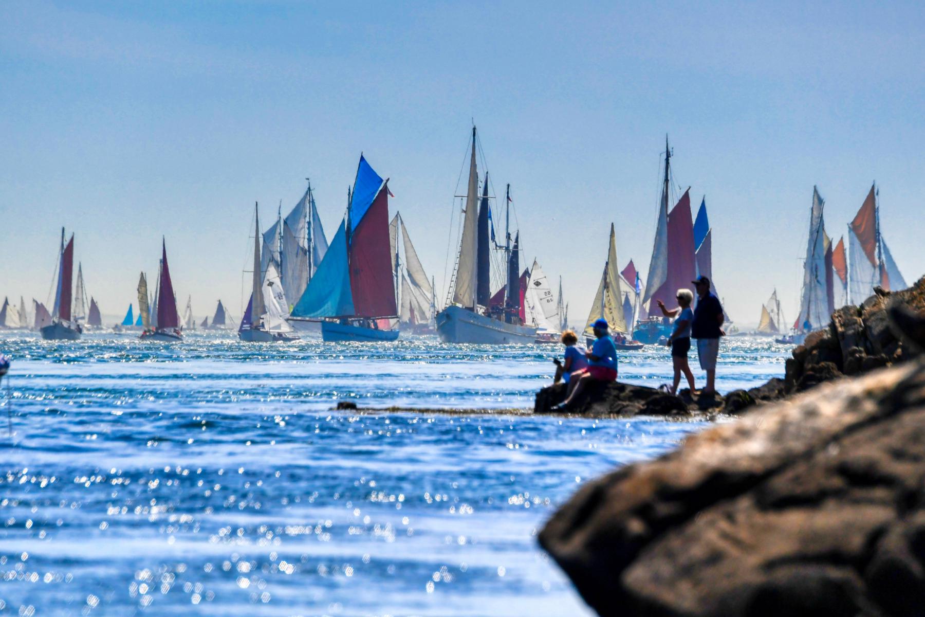 Voiliers traditionnels aux voiles colorées naviguent sur une mer bleue face à des spectateurs assis sur des rochers.