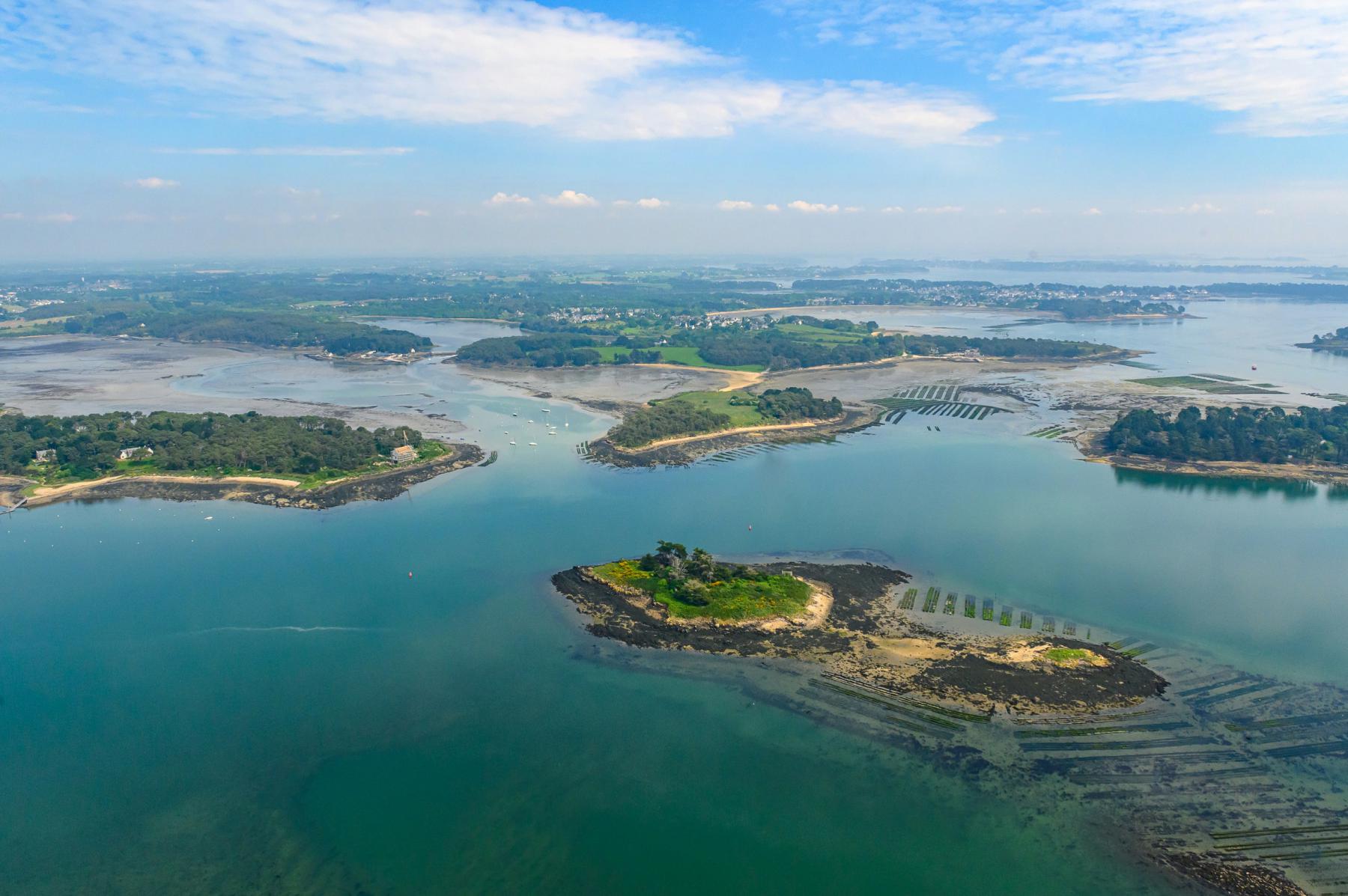 Vue aérienne sur les îles Ernic entourées d’eaux turquoise et de parcs à huîtres dans le Golfe du Morbihan sous un ciel azur.
