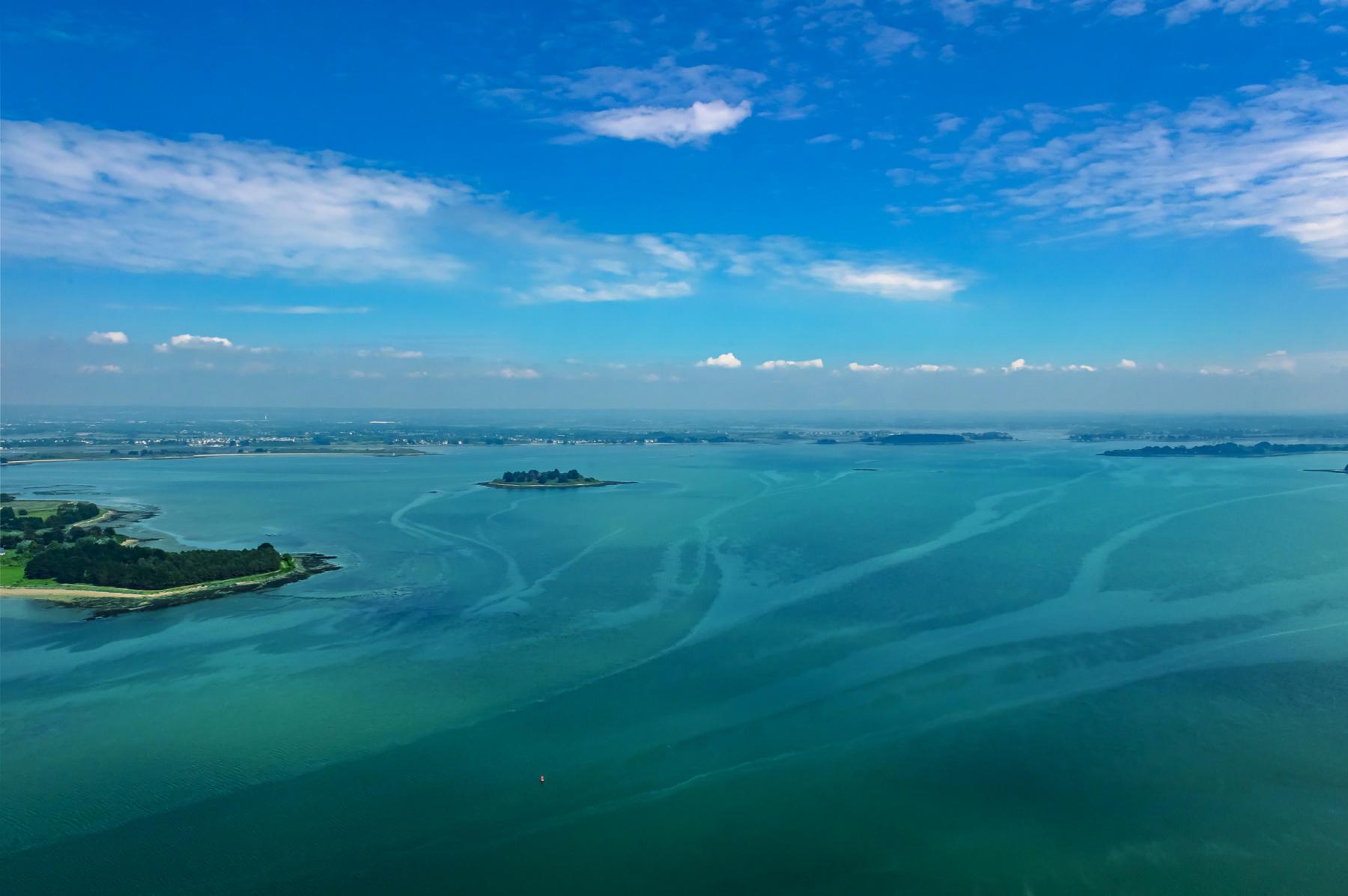 Eaux turquoise du Golfe du Morbihan entourant les îles d’Arz et de Lern sous un ciel bleu d’été parsemé de nuages.