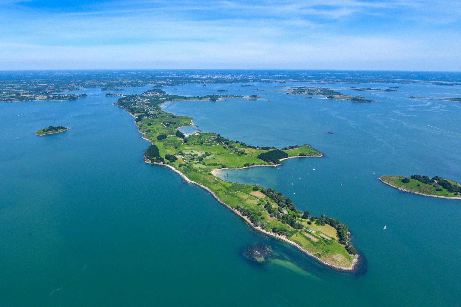 Vue aérienne panoramique de l’île aux Moines entourée des eaux bleues du Golfe du Morbihan sous un ciel clair ensoleillé.