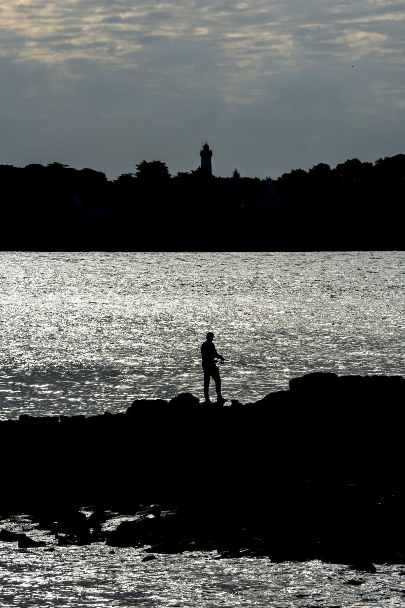 Silhouette de pêcheur sur les rochers devant la mer scintillante et le phare de Port-Navalo en arrière-plan.