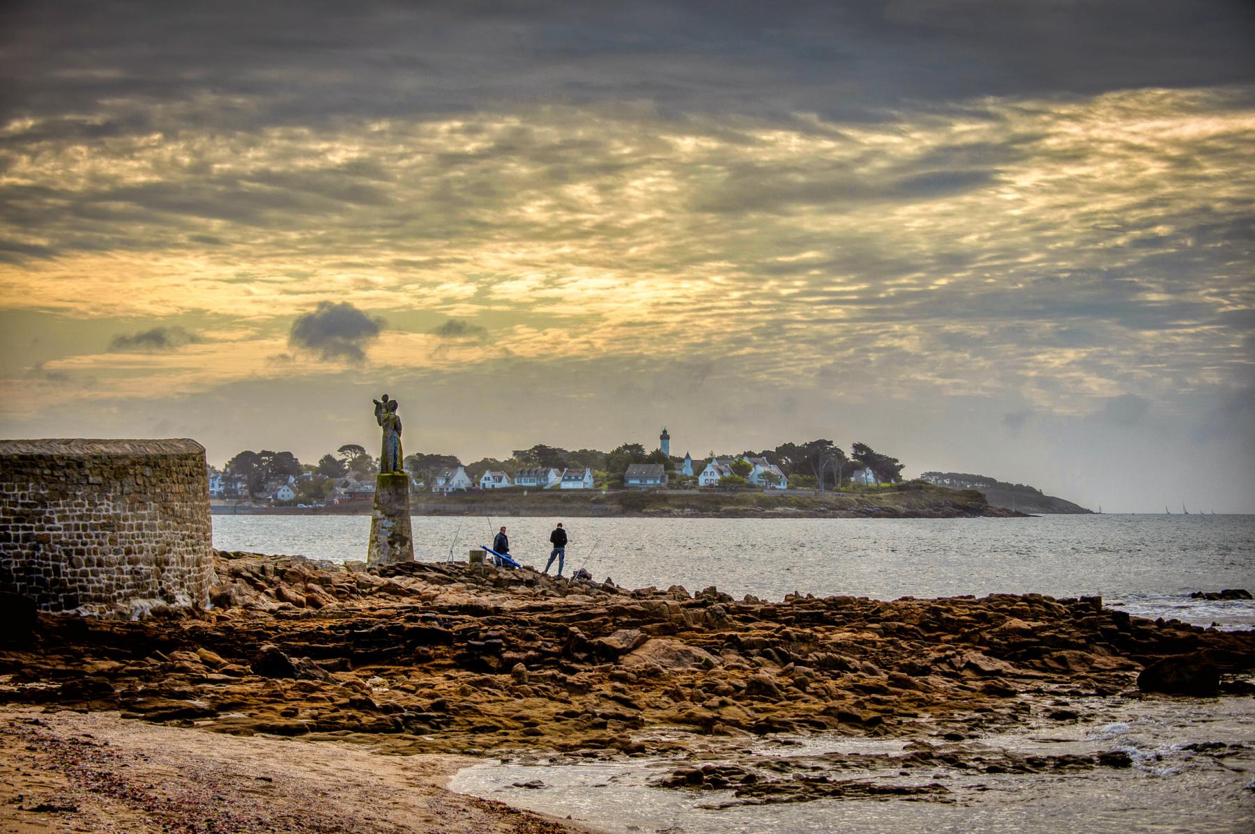 Statue de la Vierge sur un socle en pierre face à la mer à la pointe de Kerpenhir sous un ciel nuageux au crépuscule.