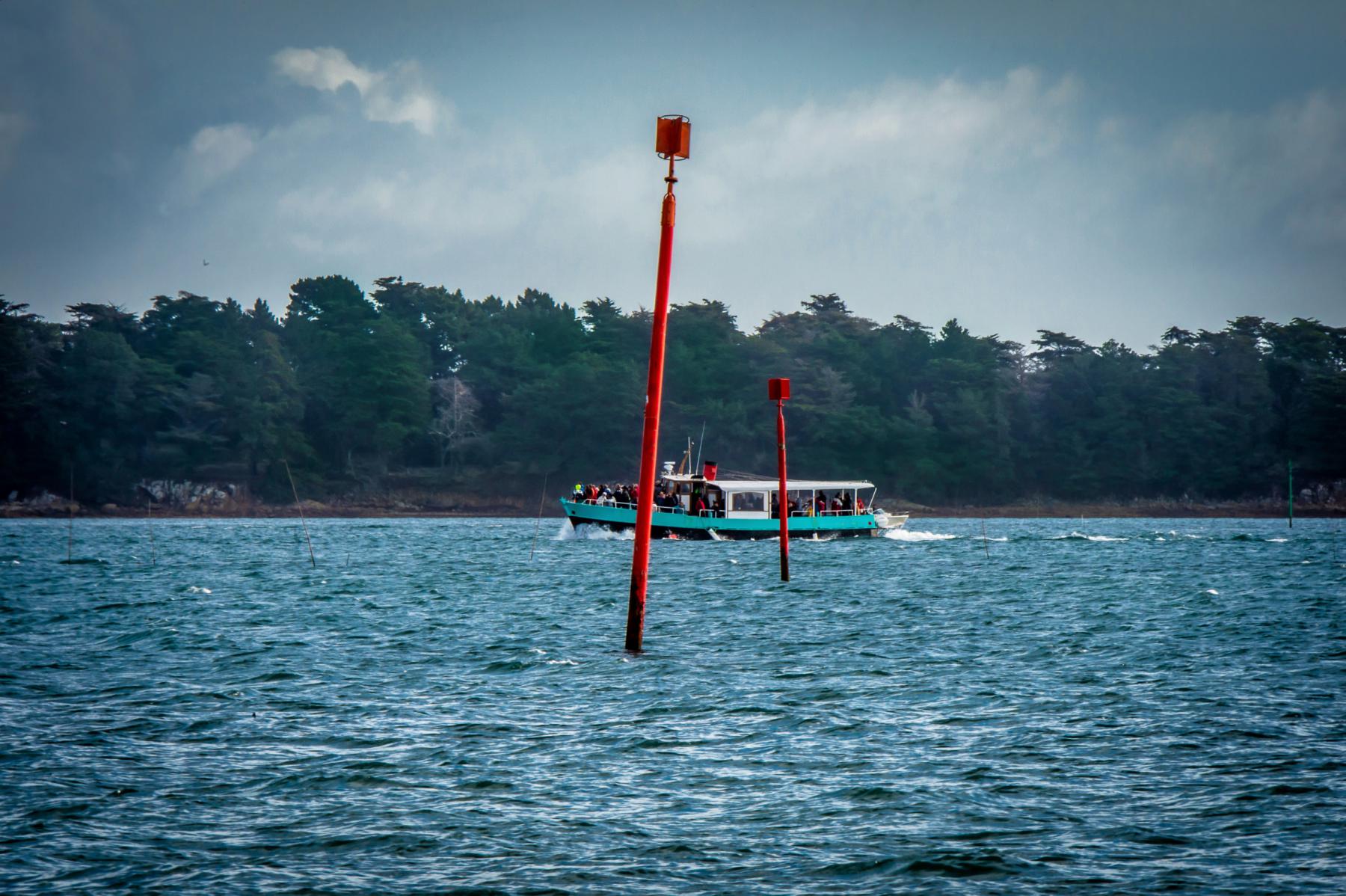 Bateau de passagers bleu et blanc naviguant sur une mer agitée entre deux balises rouges dans le Golfe du Morbihan.