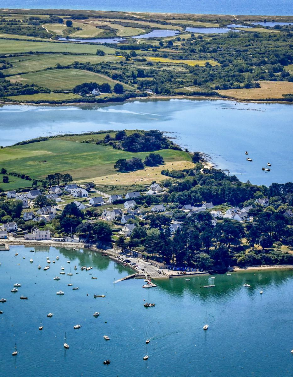Vue aérienne du petit port du Logeo avec de nombreux bateaux au mouillage sur une eau turquoise bordée de maisons et de verdure.