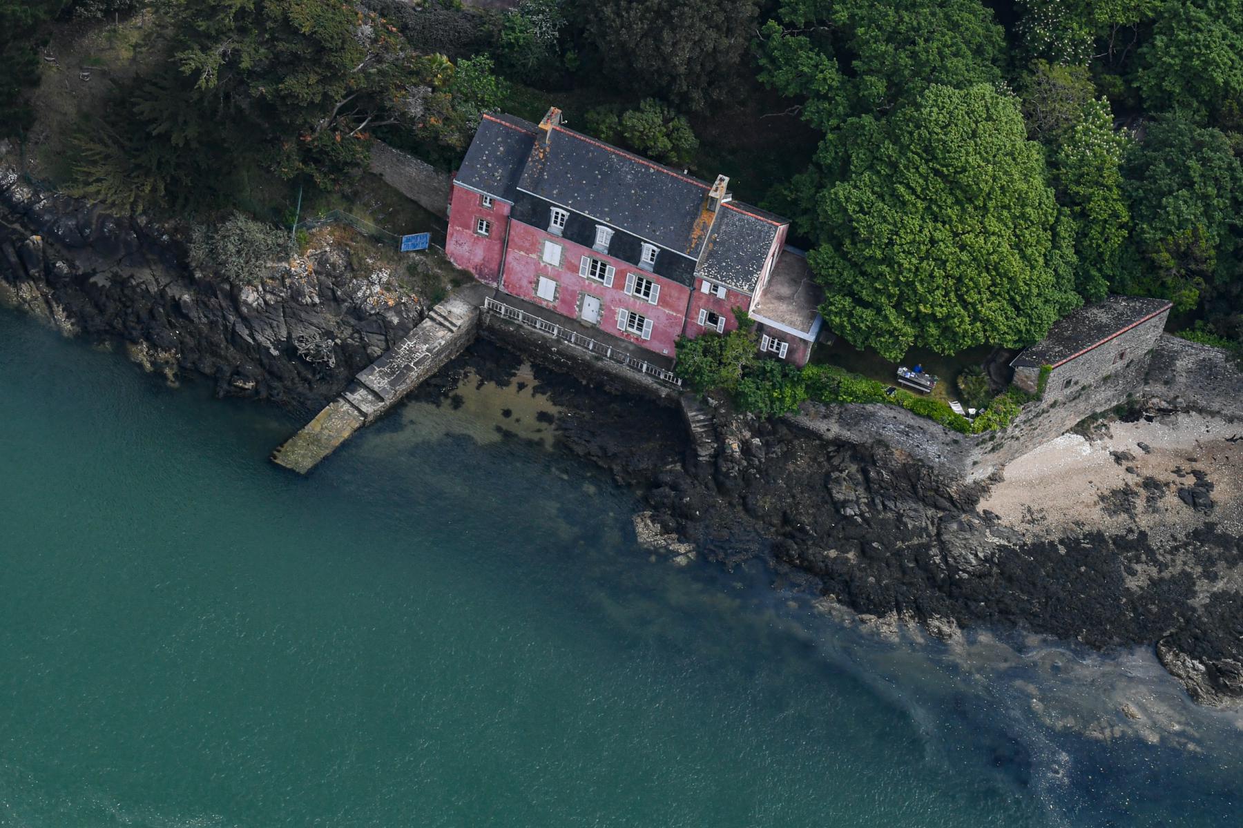 Vue aérienne d’une maison rose aux volets blancs sur une côte rocheuse bordée d’une mer turquoise et d’arbres verts.