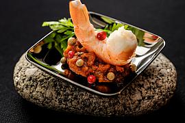 Gambas dressée sur une compotée d’épices et baies roses dans une coupelle noire posée sur un galet brut.